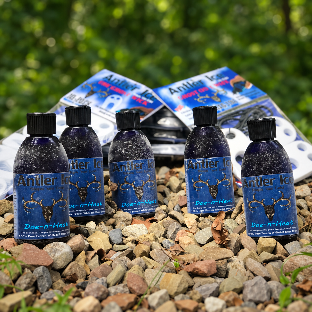 Five bottles of Antler Ice Doe-n-Heat liquid repellent are arranged on a bed of small gray and brown rocks, with two matching blue product boxes behind them. The background is a blurred green outdoor setting.