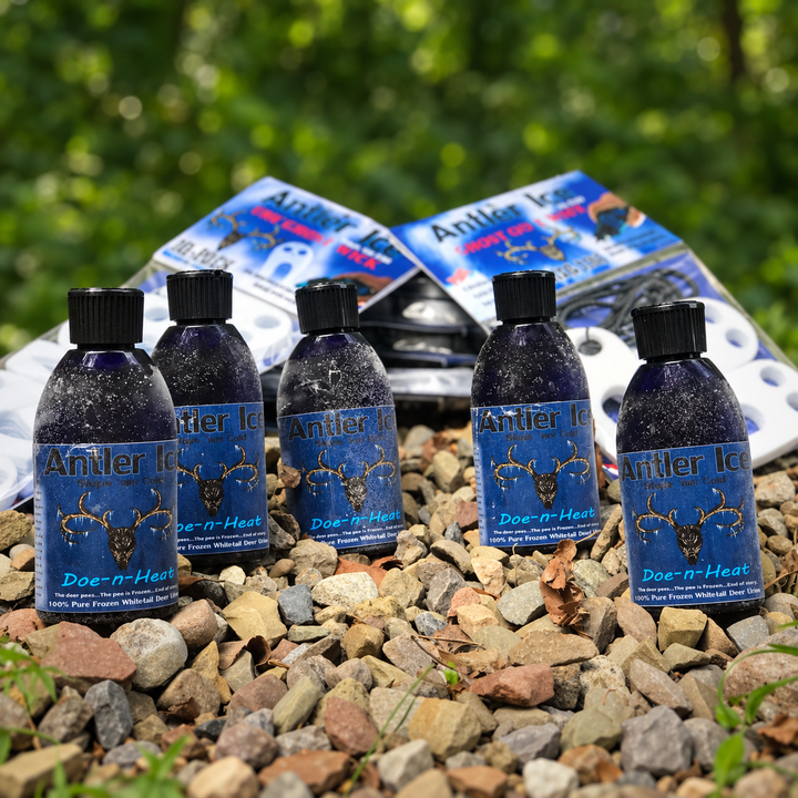 Five bottles of Antler Ice Doe-n-Heat liquid repellent are arranged on a bed of small gray and brown rocks, with two matching blue product boxes behind them. The background is a blurred green outdoor setting.