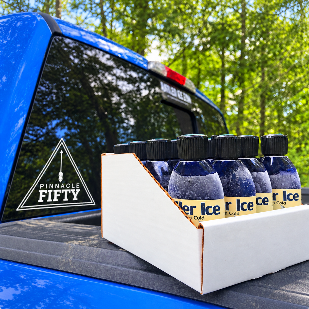 A white cardboard box filled with small dark bottles of Antler Ice frozen deer urine, branded with Pinnacle Fifty, placed on the bed of a blue truck outdoors with trees in the background.