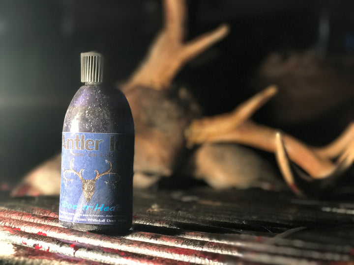 A 4 oz bottle labeled 'Doe-n-Heat Urine' with a deer silhouette, placed on a surface with deer antlers in the background.