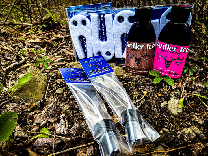 Two bottles of elk urine, one with a brown label and one with a pink label, two spray caps packaged in plastic and labeled and a 5 pack of Ghost Wicks placed on the ground in the dirt with some early spring green sprouting plants